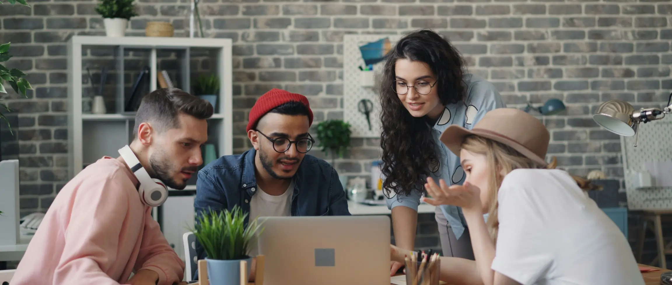 Four young adults gathered around a laptop in a modern workspace. They appear to be collaborating on a project, with a casual, creative atmosphere. The setting includes a brick wall, plants, and shelves in the background.