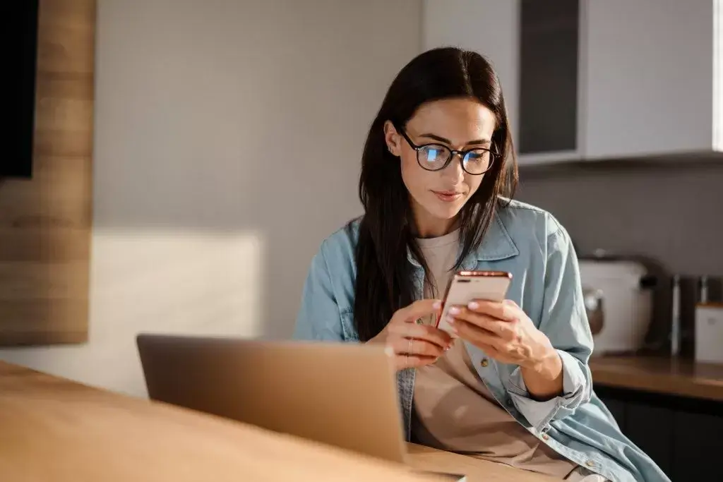 Image of a lady sat at a desk , she has a smartphone in her hand and is looking at it.