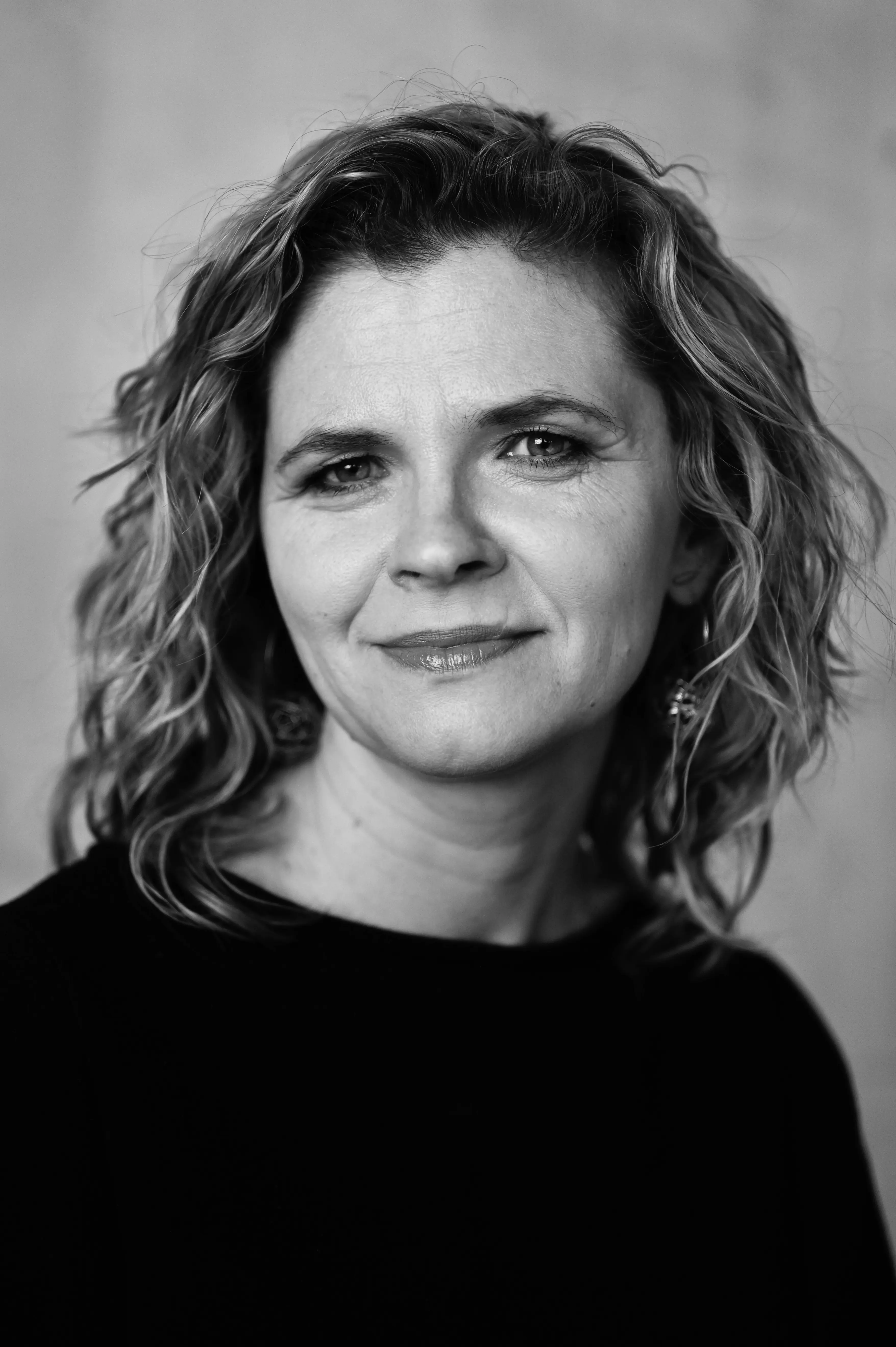 Portret czarno-biały kobiety o kręconych włosach z lekkim uśmiechem na twarzy, patrzącej prosto w obiektyw.