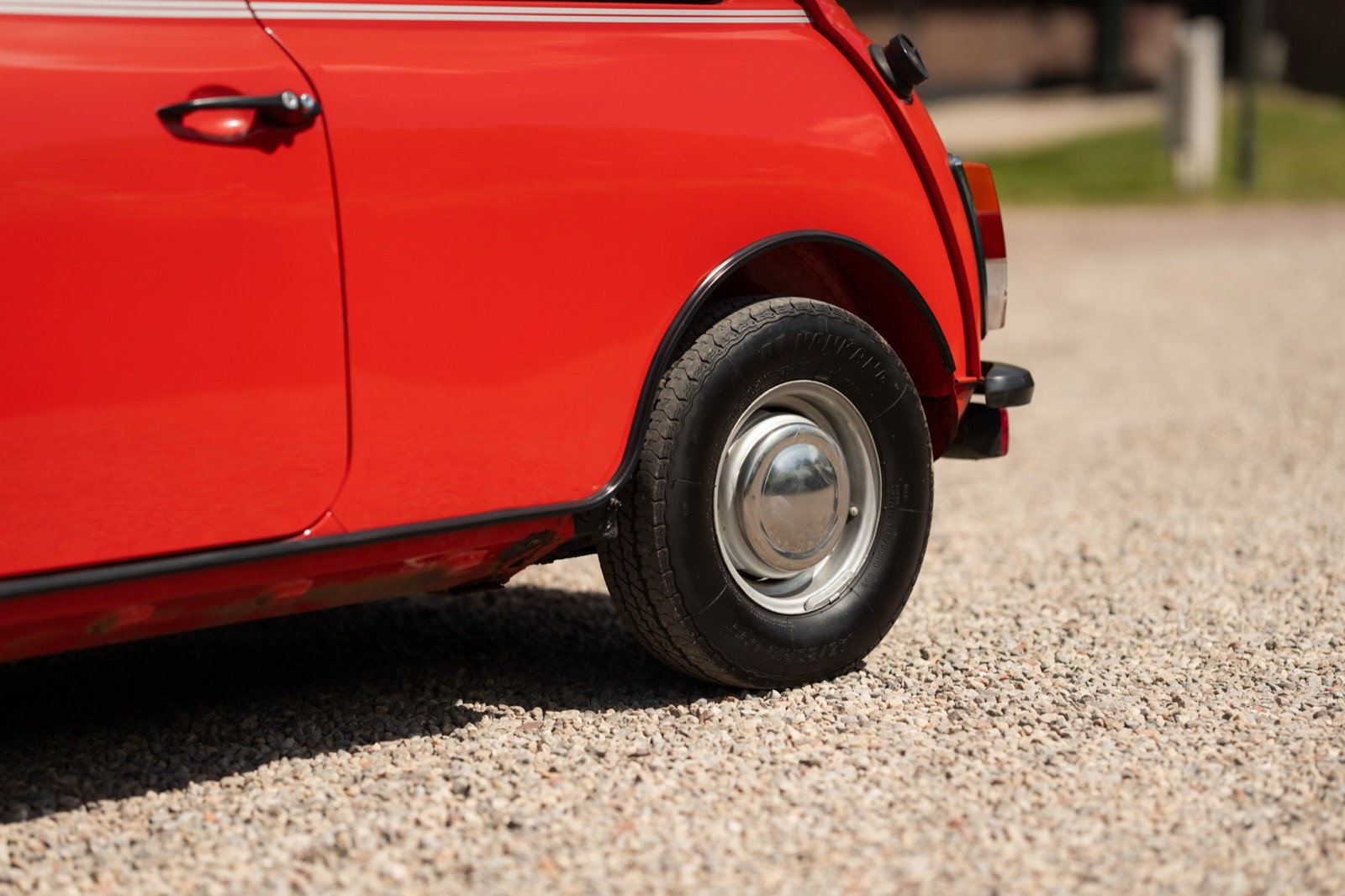 A close-up of the rear wheel of a bright red car parked on gravel, with greenery in the background.