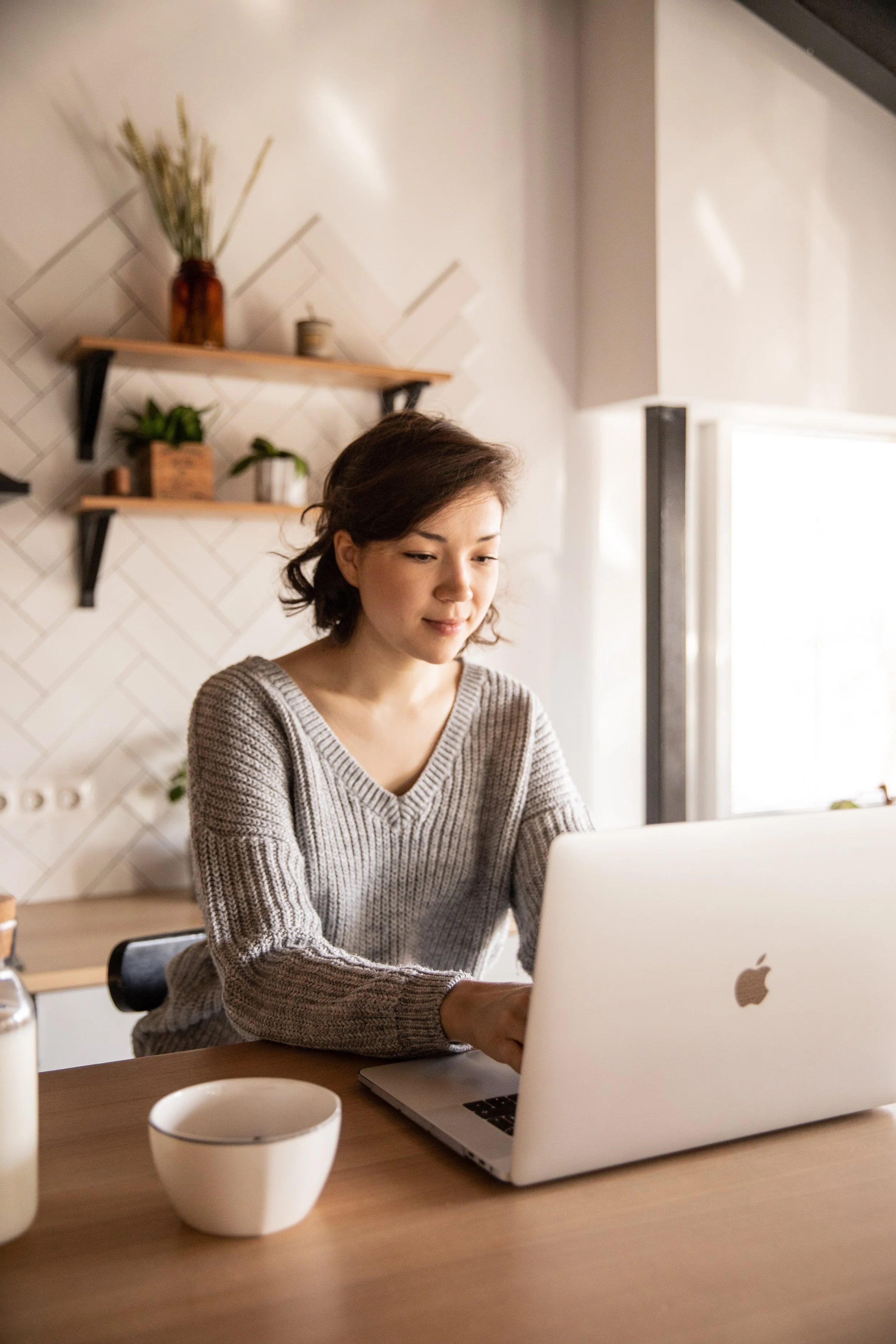 A woman sits at her kitchen counter with a cup of tea and her laptop