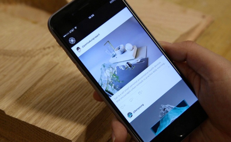 A hand holds a black smartphone displaying a social media app interface. The screen shows two posts: one from @banikunprakert and another from @beryoung. The phone rests on a light brown wooden surface.