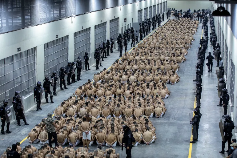 Hundreds of suspected gang members sit on the floor in a mass prison. Security officers and next to the walls.