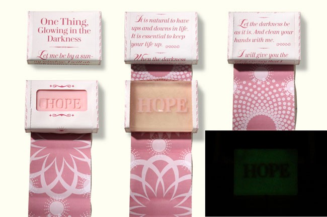 The image shows three boxes of soap, each with a pink ribbon-like design and a white label. The box on the left has a pink label with white text reading “One Thing, Glowing in the Darkness” and “Let me be by a sun-”. The center box has a beige label with white text reading “It is natural to have ups and downs in life. It is essential to keep your life up.”. The box on the right has a pink label with white text reading “Let the darkness be as it is. And clean your hands with me.”. The rightmost box has a green label with white text reading “I will give you the”. The boxes are displayed against a white background.