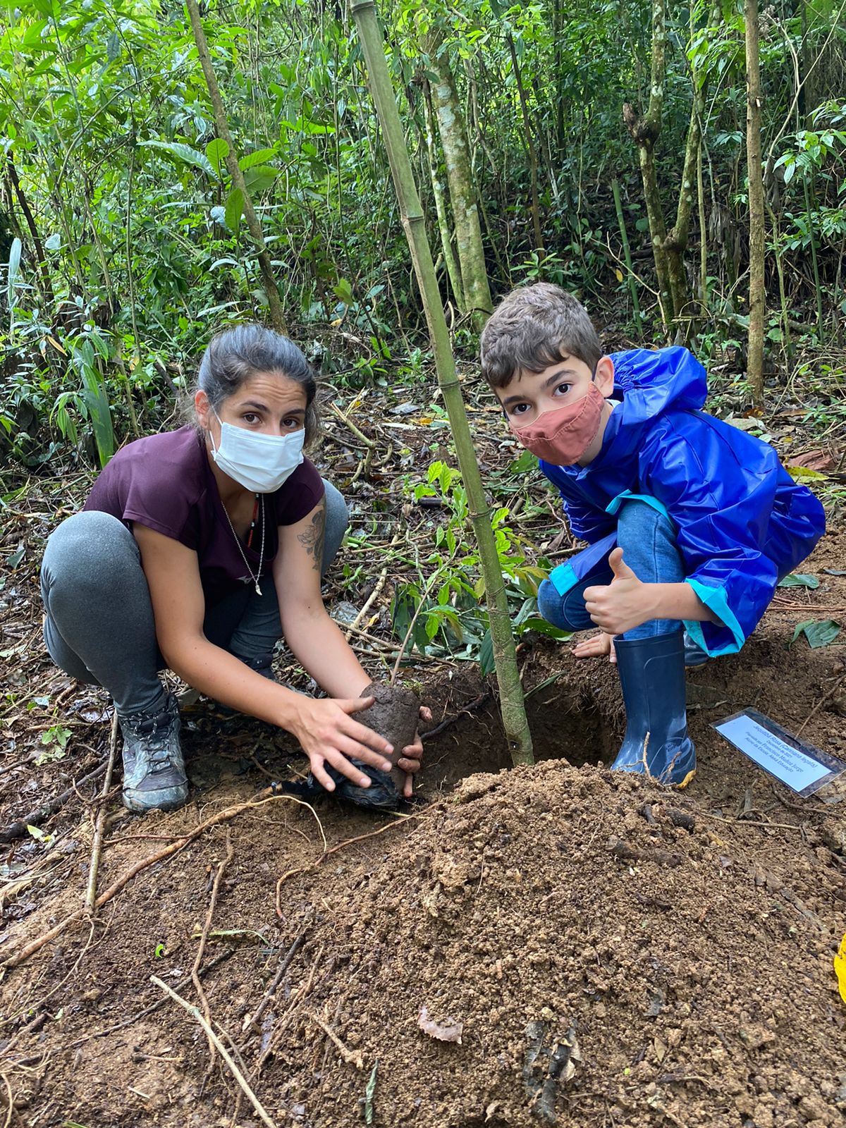 Mulher e criança de máscara plantam muda de árvore em cova aberta no meio da mata
