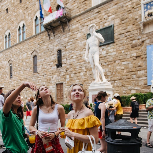 Tři ženy se dívají vzhůru před velkou kamennou budovou se sochou Davida venku. V blízkosti jsou další lidé a odpadkový koš.