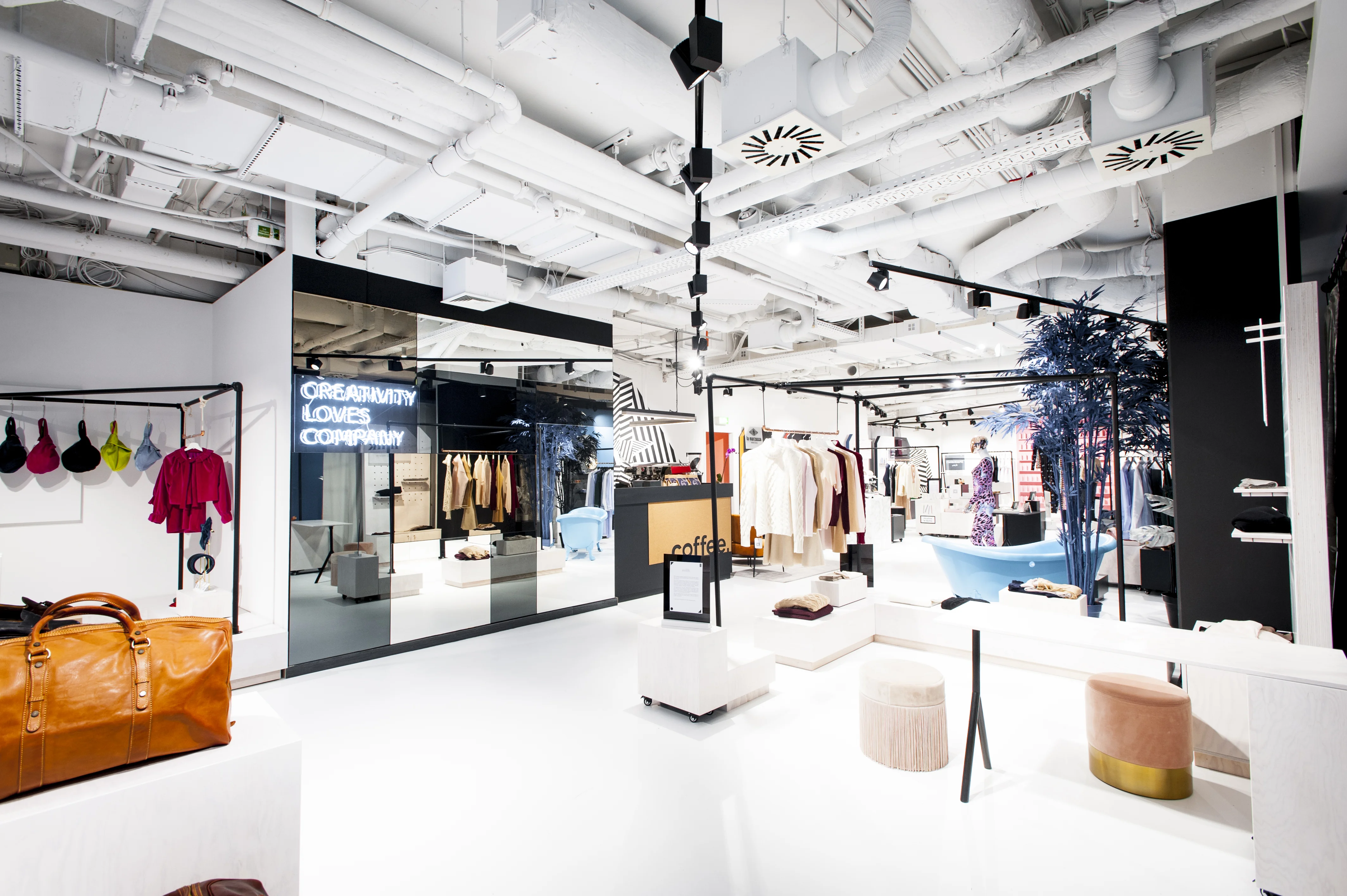 Overview of the FREIRAUM SHOP concept store with white exposed-services ceiling, clothing racks, a leather weekender on a plinth, velvet poufs and a Creativity Loves Company neon in a mirrored box.