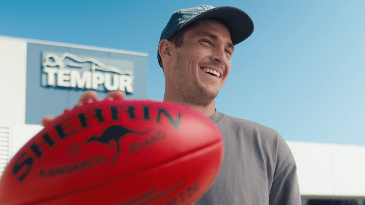 A young man wearing a cap and light-colored shirt holds up a red boxing glove, with a blue sky visible in the background