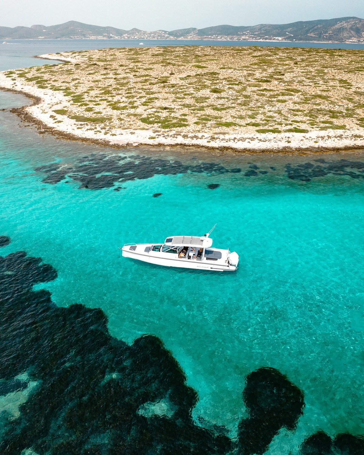 White Axopar 37 yacht anchored in turquoise waters near a small uninhabited island with sparse vegetation in the Cyclades.