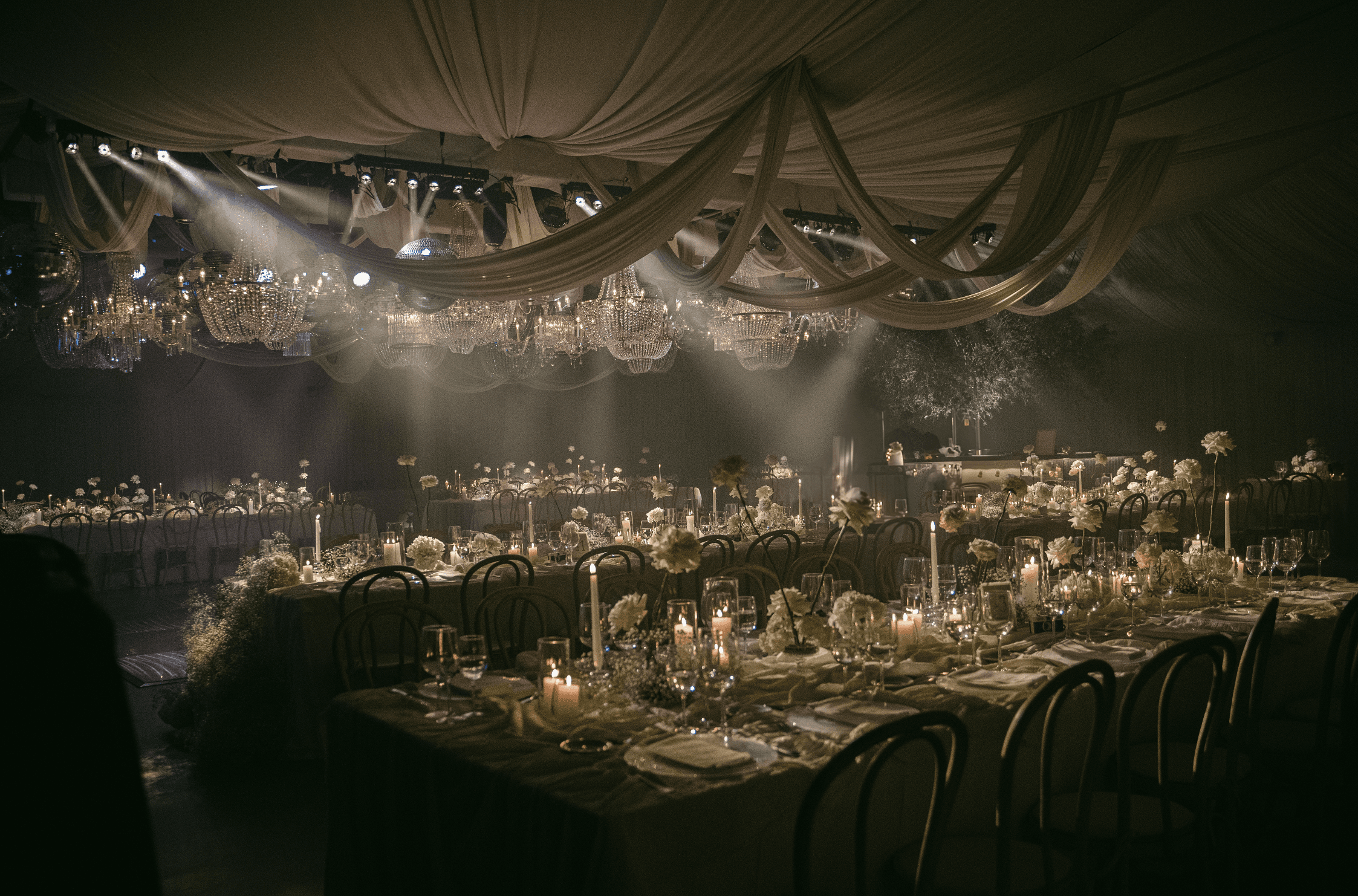 Salón de eventos con iluminación baja, arañas de cristal colgantes, telas etéreas y mesas largas decoradas.