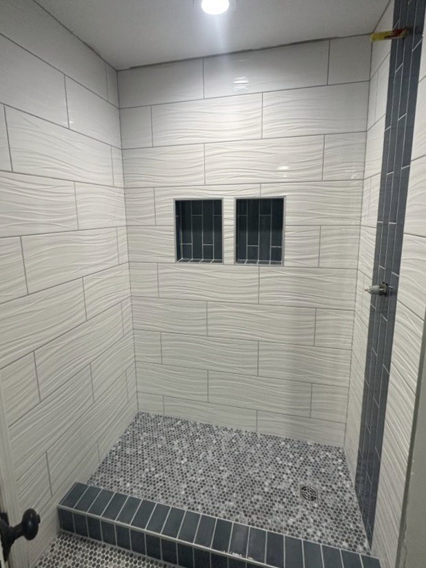 Modern shower featuring textured white wavy wall tiles, two built-in niches with dark grey subway tile inserts, a vertical accent stripe of dark grey subway tiles, and a multi-toned pebble mosaic shower floor.
