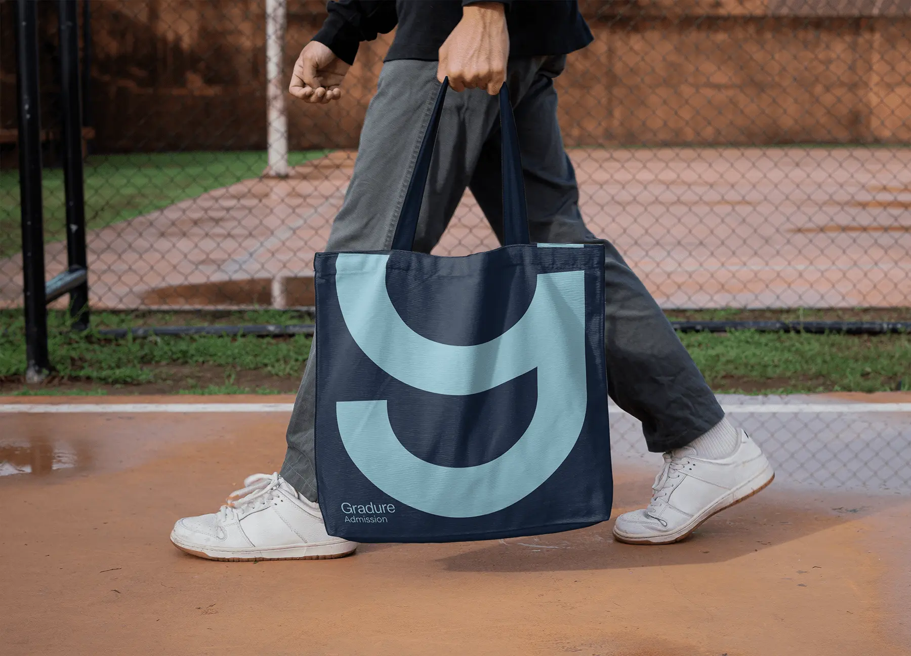 Person walking outdoors carrying a navy-blue Gradure Admission tote bag featuring a large light-blue geometric ‘G’ symbol as the main graphic.