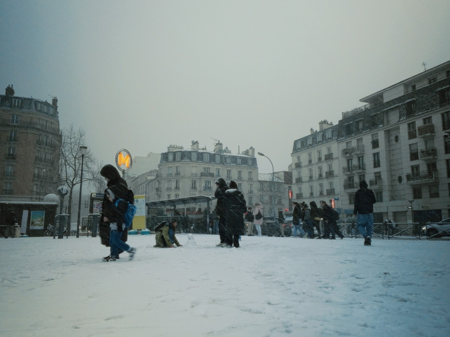 Place enneigée à Paris, panneau "M" de métro, piétons en hiver, enfant construisant un bonhomme de neige, ciel nuageux.