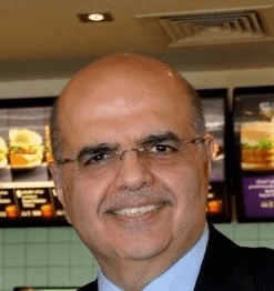 A smiling man with short brown hair in a dark blazer and white shirt against a light gray background. His expression conveys warmth and friendliness.