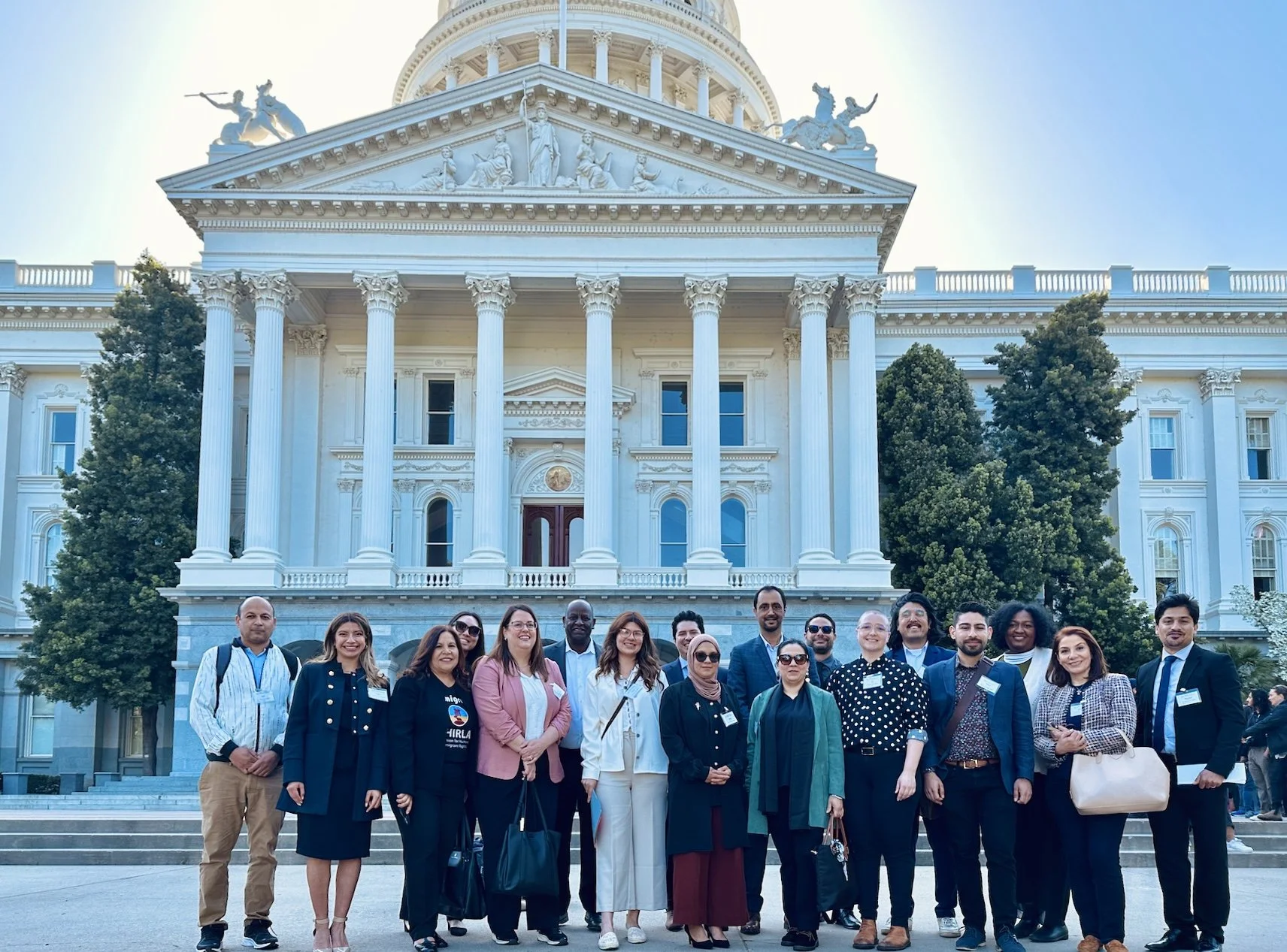 Um grupo diversificado de pessoas está em frente à majestosa arquitetura neoclássica do edifício do Capitólio branco, destacando suas grandes colunas e esculturas intrincadas em um dia ensolarado.