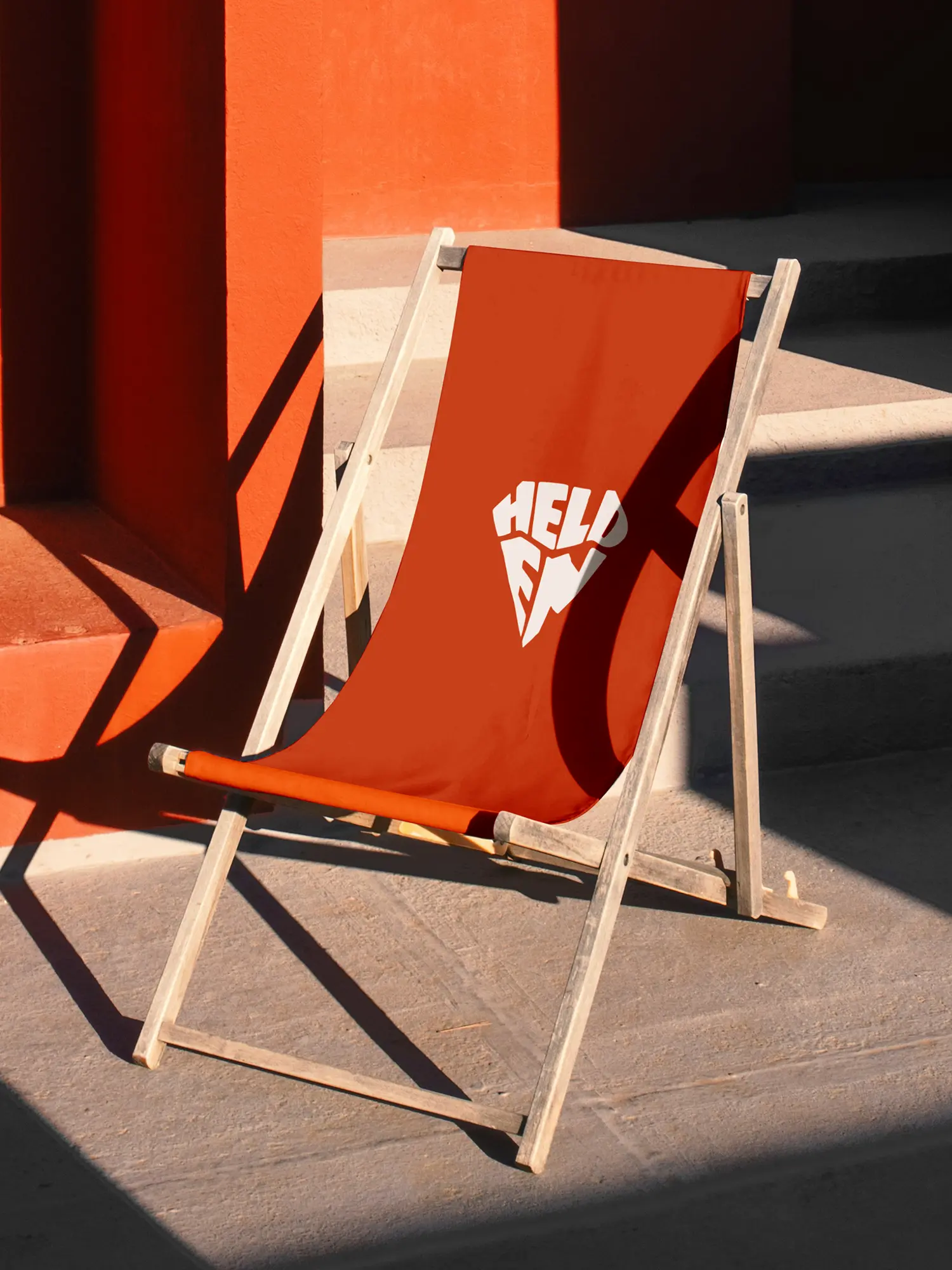 Liegestuhl mit rotem Stoff und weißem Helden-Logo vor orangefarbener Wand im Sonnenlicht.