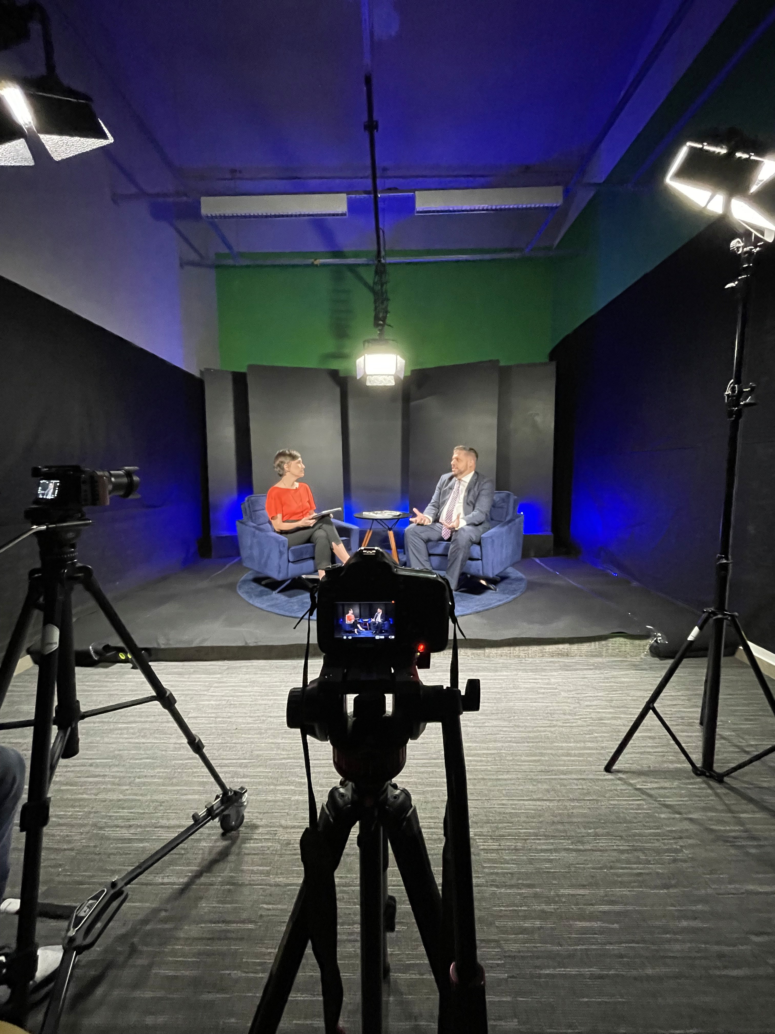 A smiling young man with crossed arms, wearing a plaid shirt and white t-shirt, poses against a dark background.