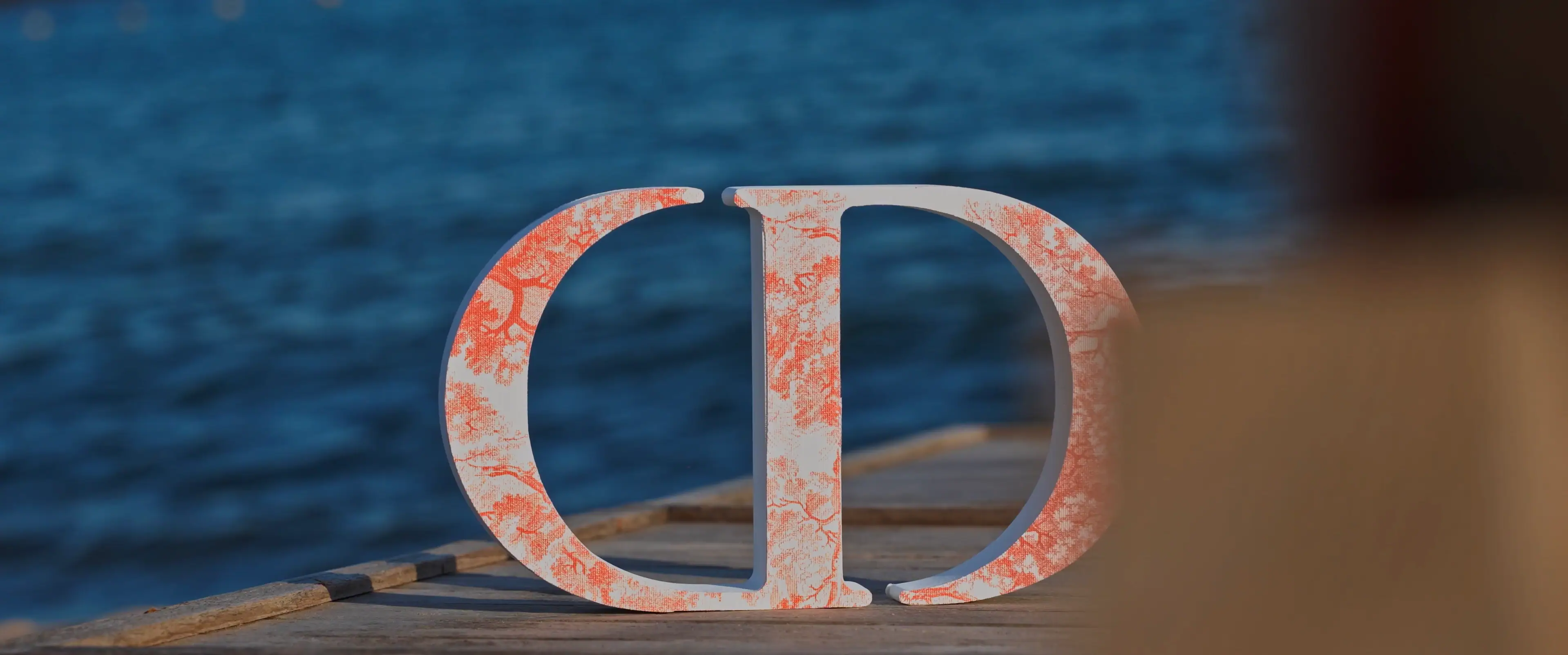 A large, ornate, coral-patterned letter 'CD' stands upright on a wooden pier, with the deep blue ocean in the background.