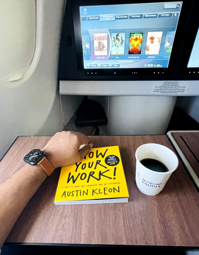 A hand with Apple watch on book titled "Show Your Work!" by Austin Kleon, cup of coffee is next to it, set in an airplane