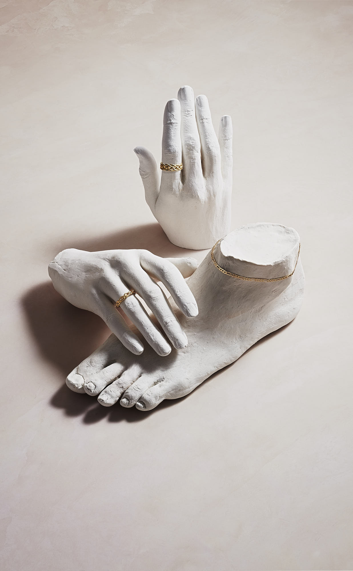 White plaster hands and foot wearing gold jewelry on light background.