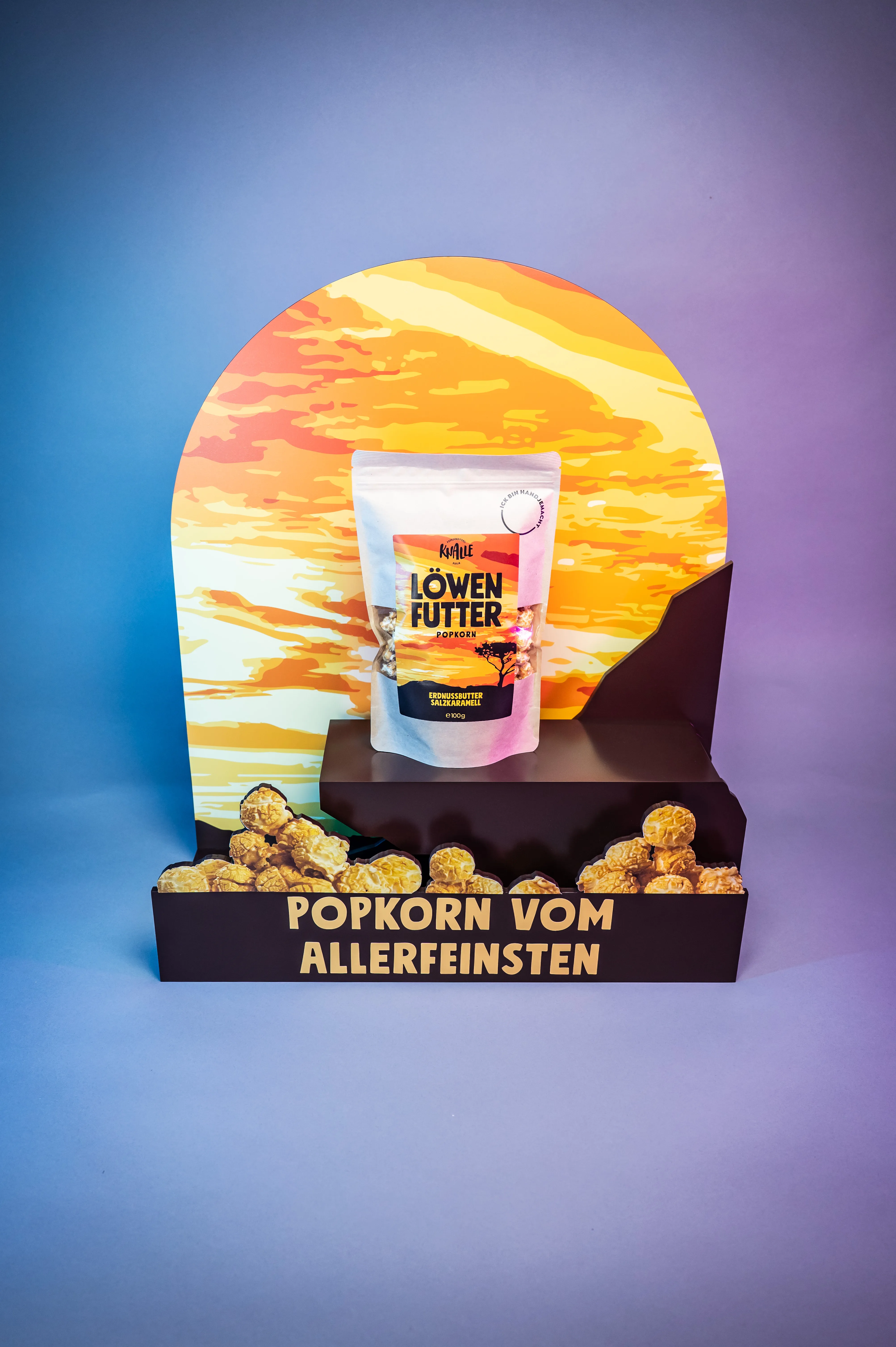 KNALLE point-of-sale display with a sunset arch backdrop, a Löwenfutter popcorn bag on a podium and the slogan Popkorn vom Allerfeinsten over cut-out popcorn.
