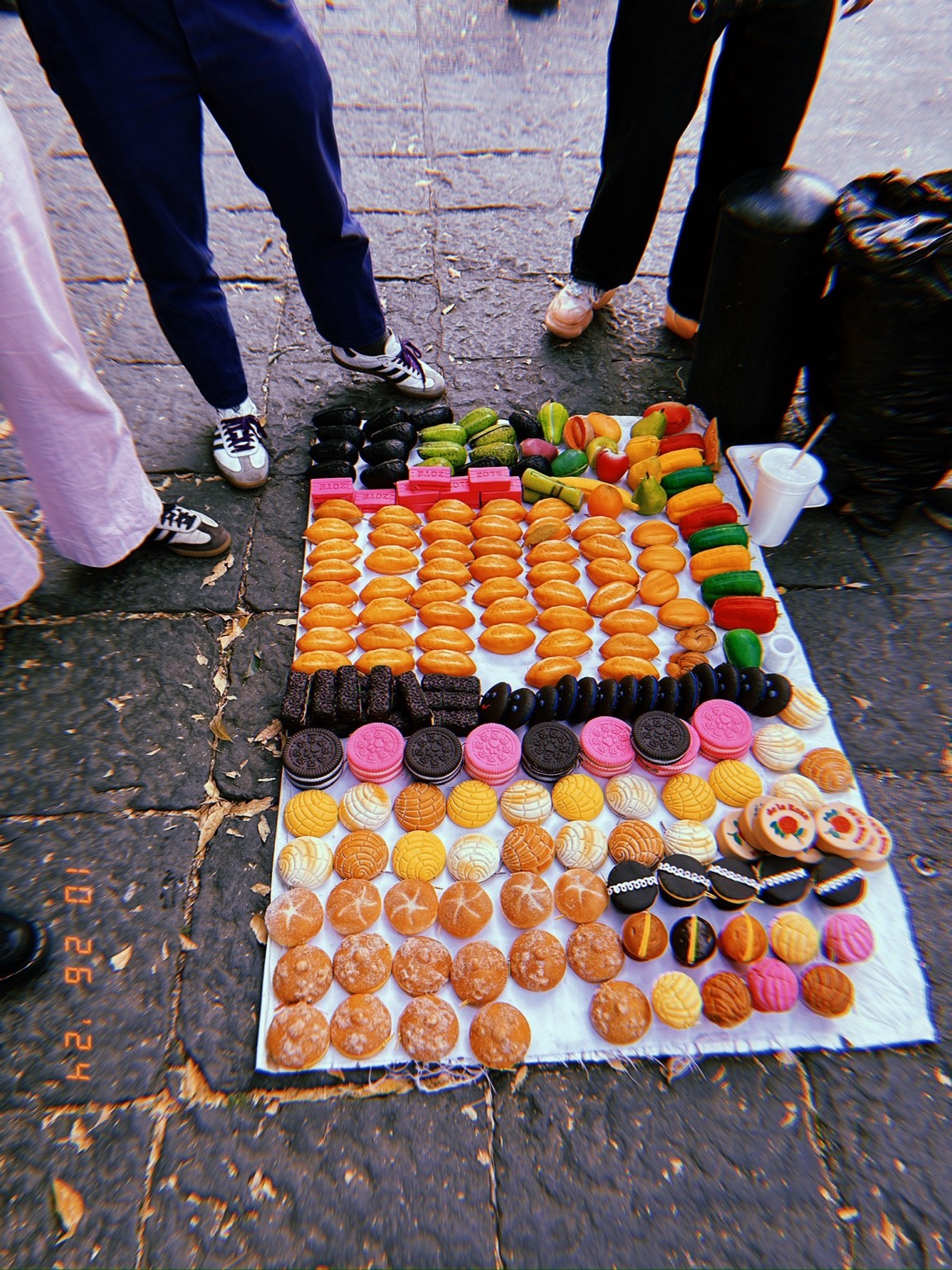 Rows of colorful miniature breads, cookies, fruit, and vegetables arranged on a white cloth on the pavement.