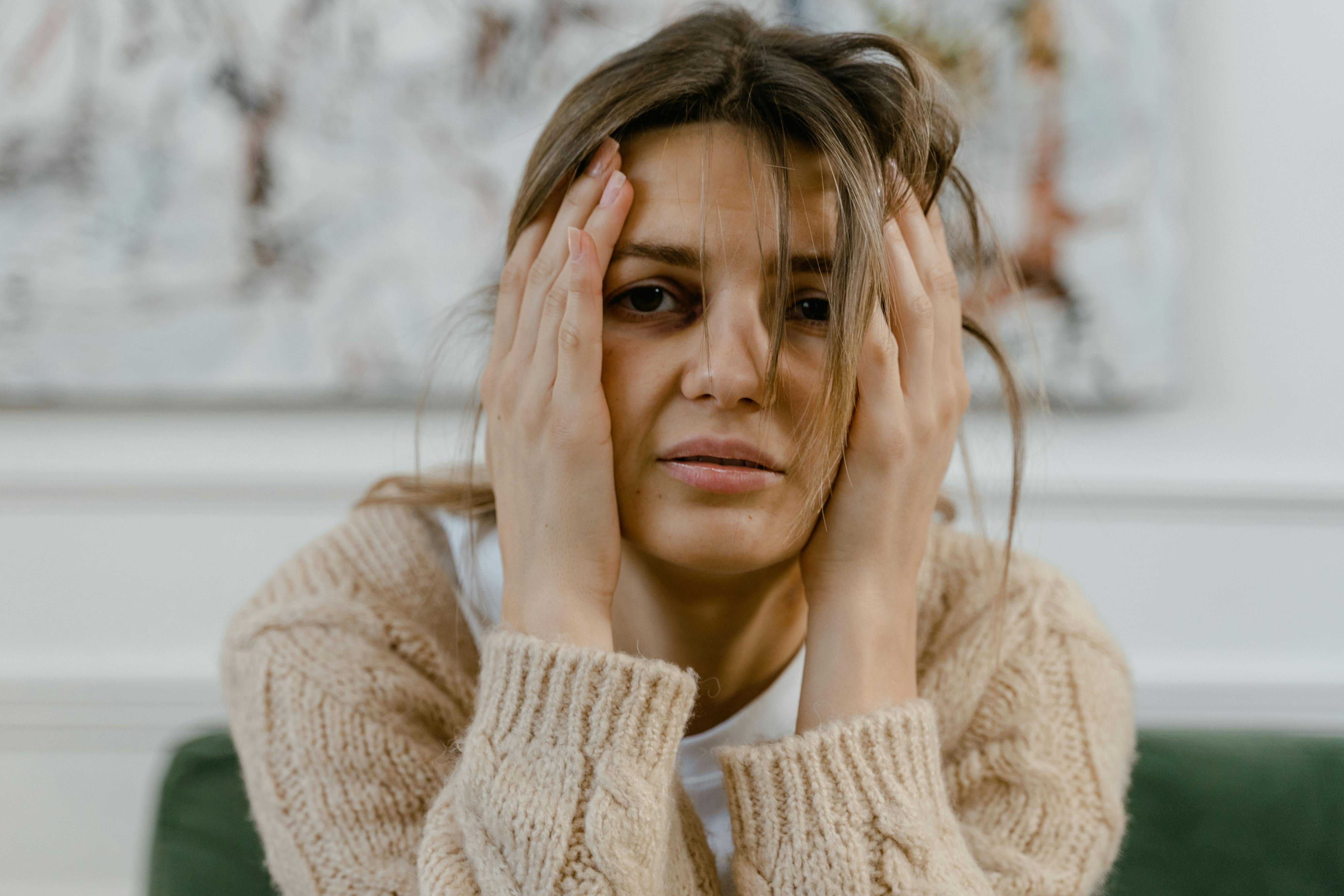 A young woman with disheveled hair and a distressed expression holds her face in her hands, conveying signs of emotional exhaustion, anxiety, or stress