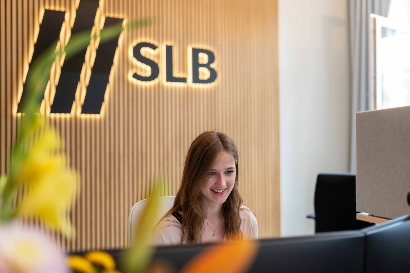 Rezeption von SLB: Freundliche Mitarbeiterin sitzt lächelnd hinter dem Empfangstresen, im Hintergrund das SLB-Logo vor einer hellen Holz- und Fensterfront.