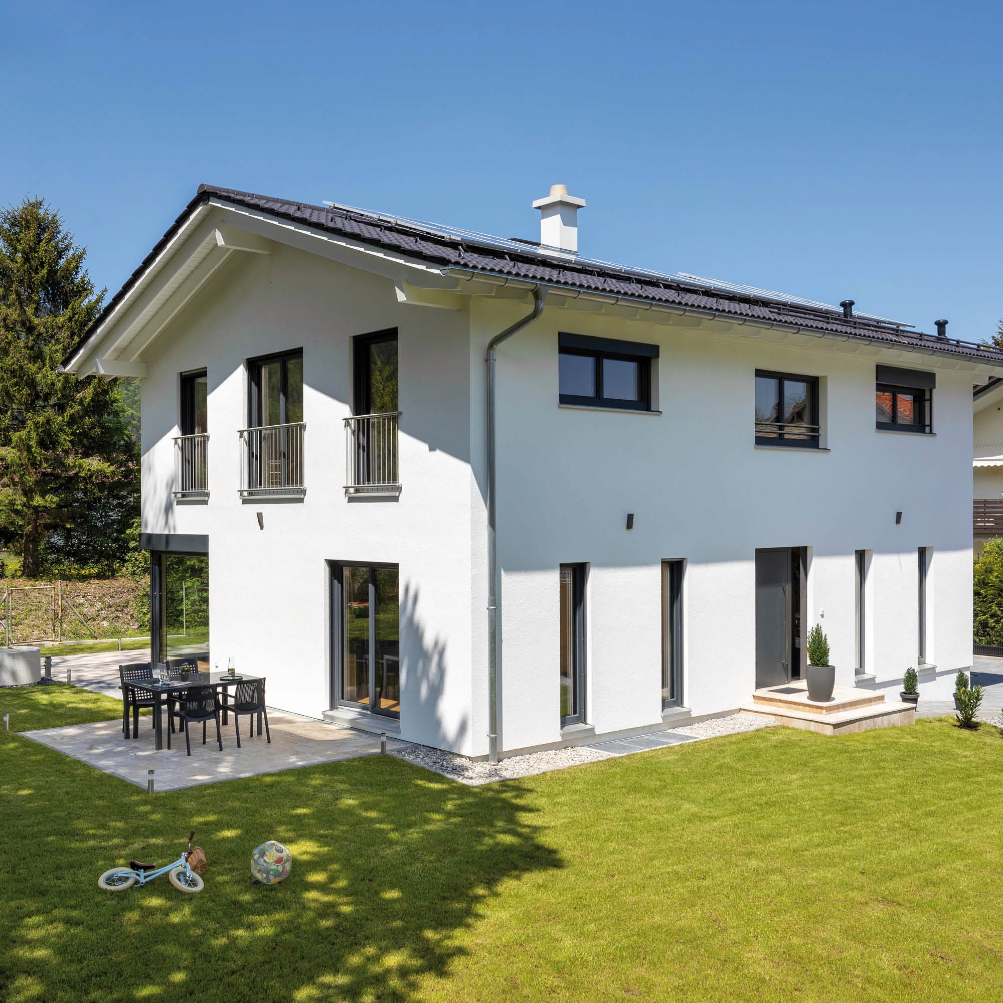 Modernes Einfamilienhaus mit weißer Fassade, dunklen Fenstern, Pedra-Terrasse und gepflegtem Garten.