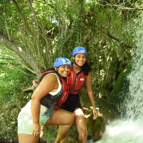 Två personer med hjälmar och flytvästar ler, stående nära ett vattenfall med frodig grönska i bakgrunden.