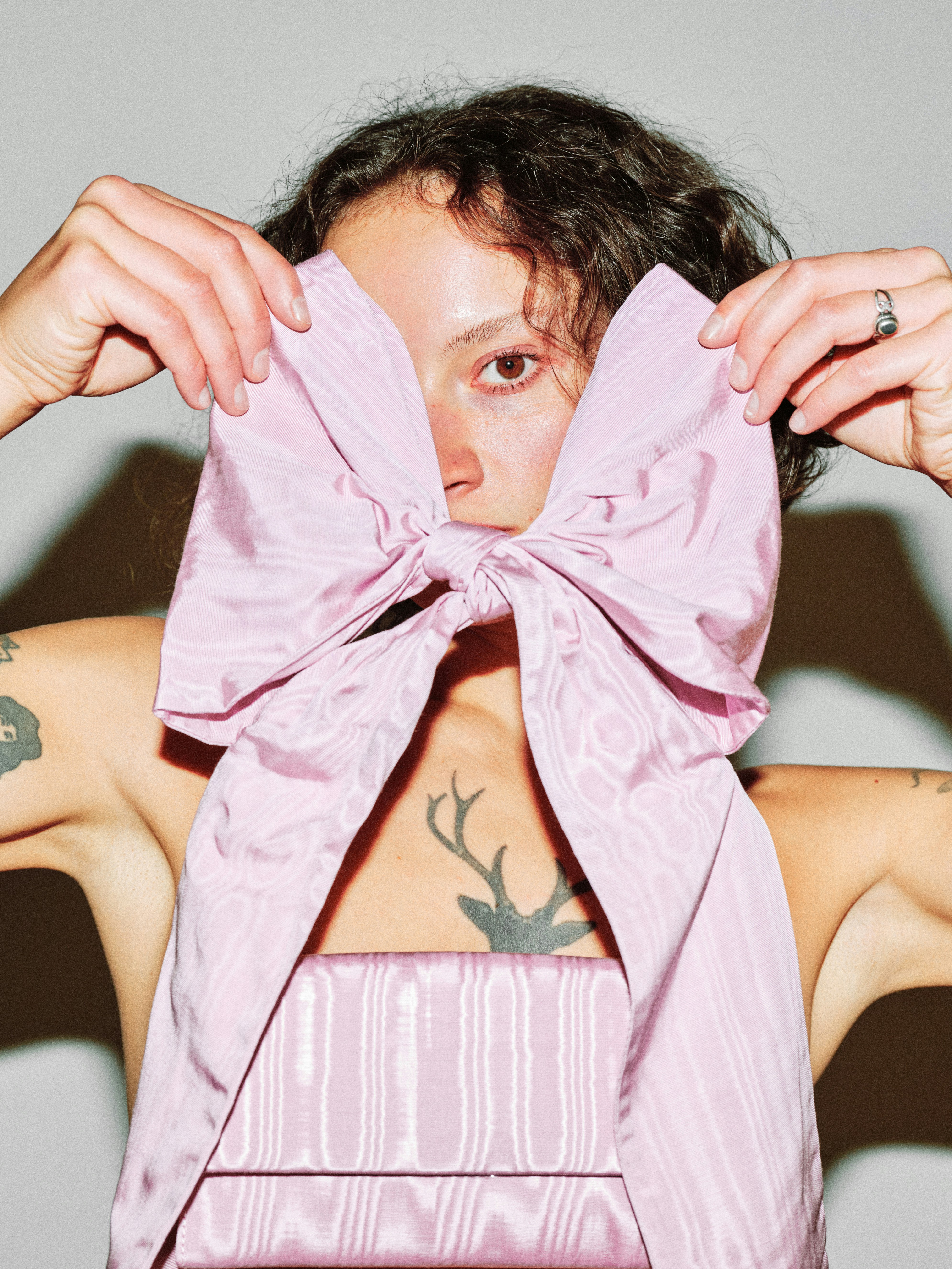 Editorial fashion portrait of a curly-haired model holding a large pink satin bow in front of her face, visible eye peering through, tattoos on arms