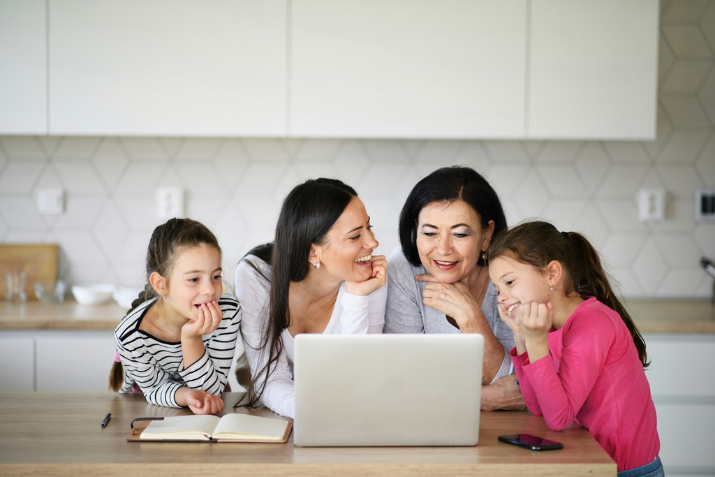 A multigenerational family of four enjoys a moment together in a modern kitchen, smiling and looking at a laptop screen. The group includes a grandmother, mother, and two young girls, highlighting the joy of shared experiences and the use of technology to connect and engage with each other.