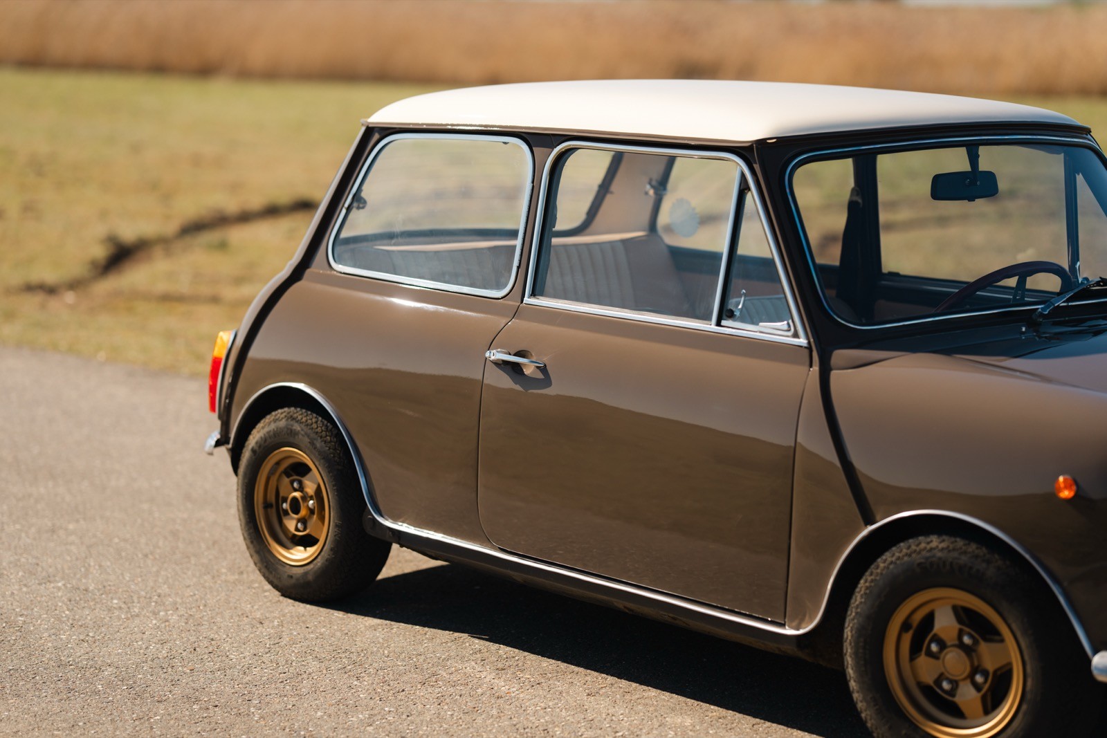 A classic brown car with gold wheels parked on a road, surrounded by grassy fields.