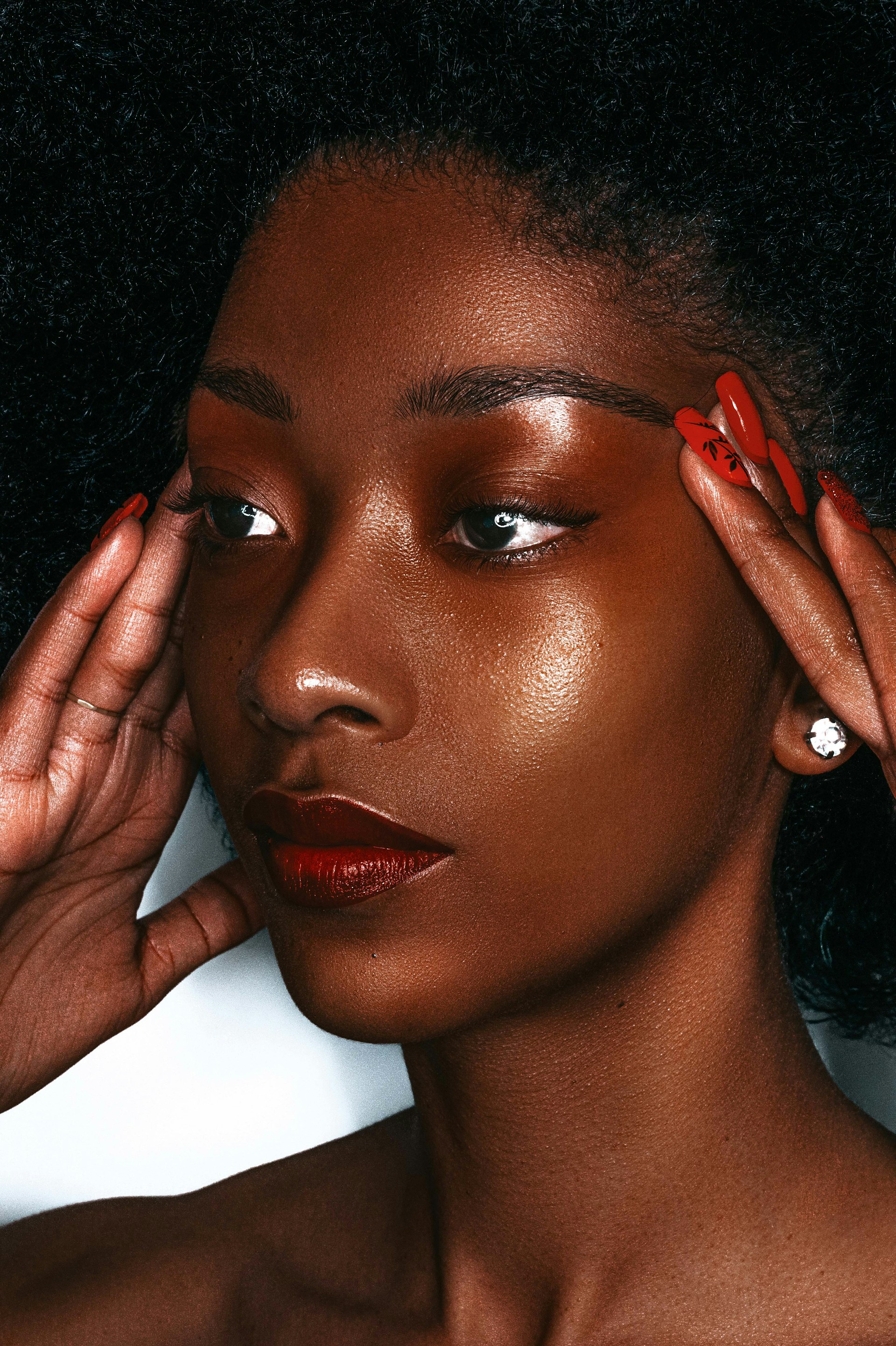 A close-up portrait of a Black woman with radiant, glowing skin and dark red lipstick, posing with her hands near her temples to highlight her red manicured nails.