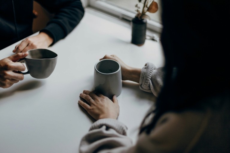 Two people sitting across a table holding mugs in a supportive conversation, representing incident support and professional guidance for healthcare workers