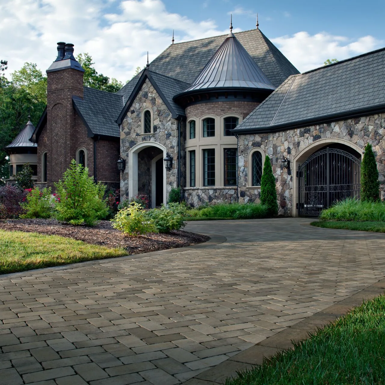 A complete property setting Dark paver driveway leading toward a large stone home with a turret and planted borders.