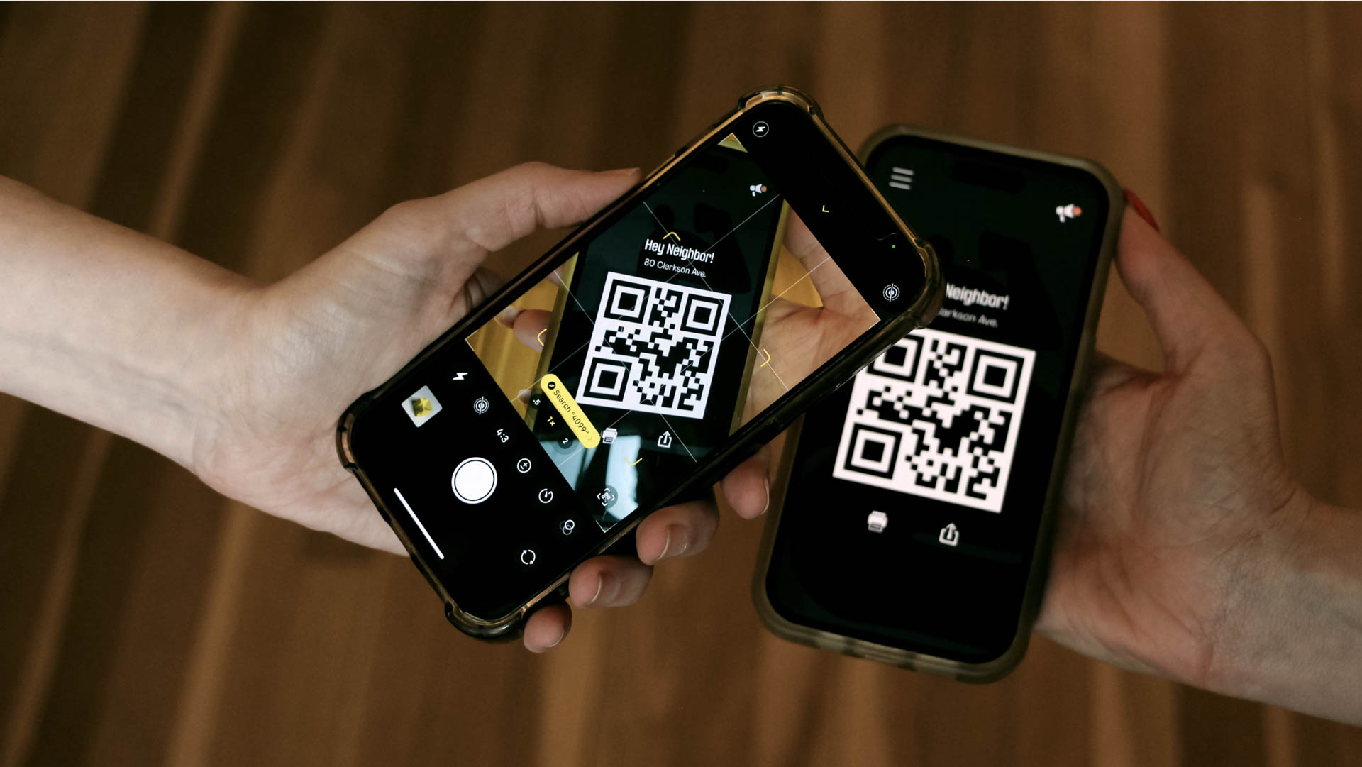 Two hands hold two smartphones displaying QR code scanning interfaces. The phone on the left shows a yellow sticker on its screen, while the phone on the right displays “Neighbor!”, both with QR codes visible on their screens. The background is a blurred wooden surface.