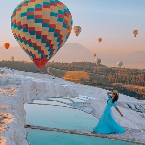 Isang babae sa asul na damit ang nakatayo sa tabi ng mga batong terrace na pool habang may makukulay na hot air balloon na lumilipad sa isang magandang tanawin ng paglubog ng araw.