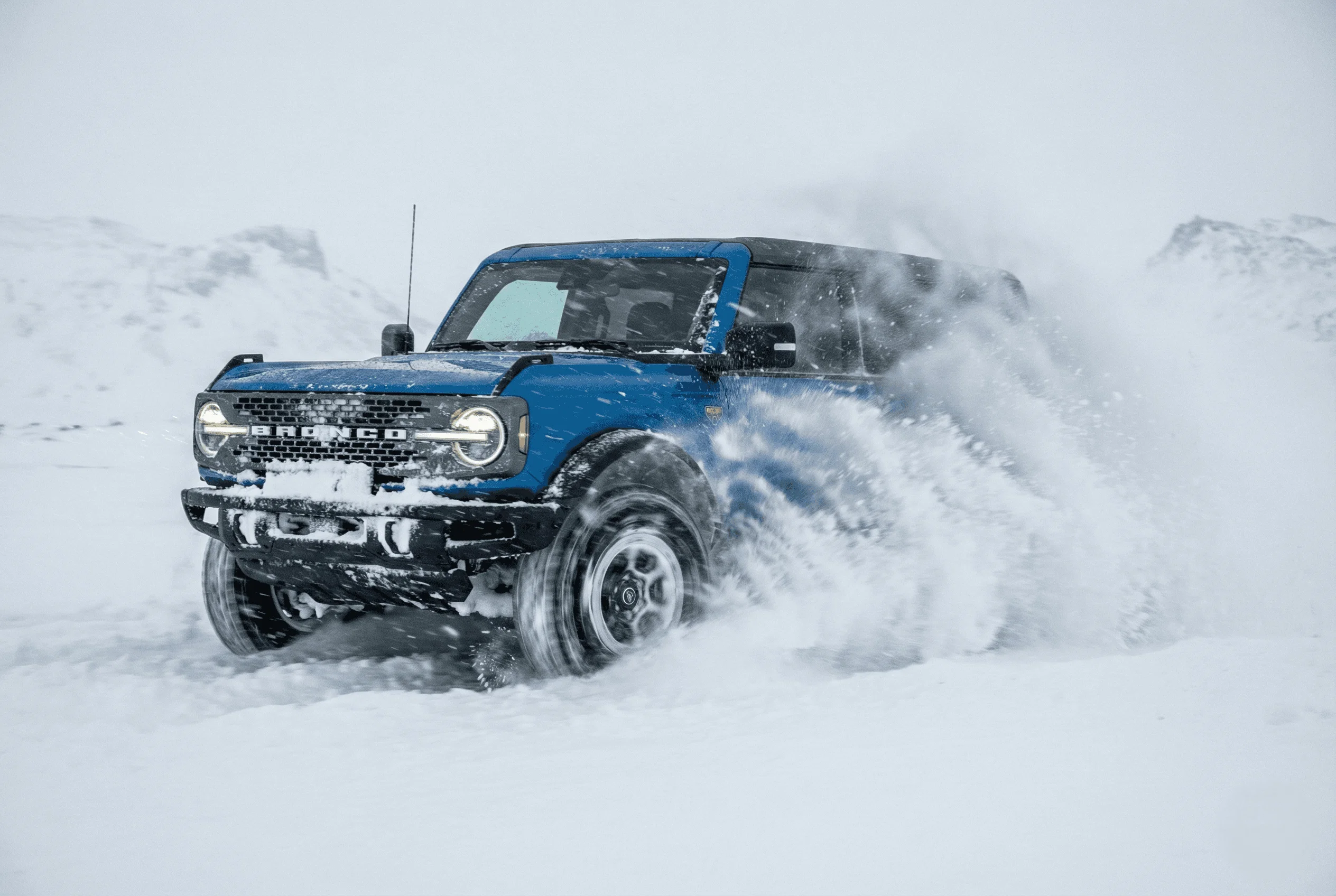 Blue Ford Bronco driving fast through deep snow, kicking up a massive cloud of white powder.