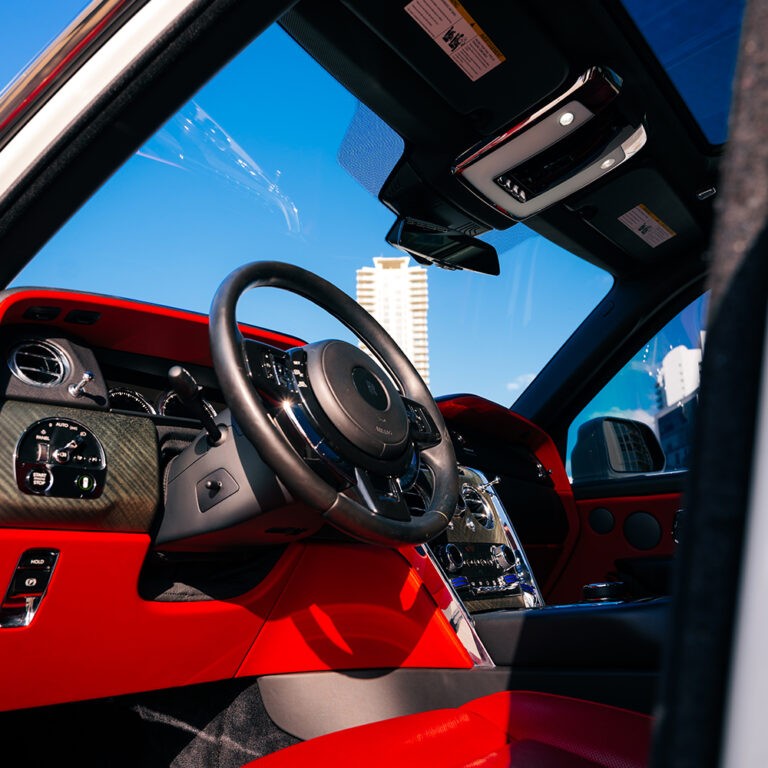 Driver's cockpit of white Rolls-Royce Cullinan, vibrant red leather interior, bespoke luxury car rental.