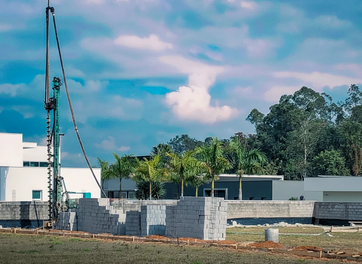 Máquina perfuratriz de grande porte em campo aberto, com blocos de concreto branco empilhados no primeiro plano. Ao fundo, um edifício moderno branco, coqueiros e uma área de mata sob um céu azul claro com nuvens brancas.