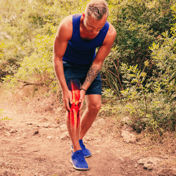 Man standing on a hiking trail reaching down and grasping his knee dressed in blue tank and shorts, with xray image over leg showing knee pain