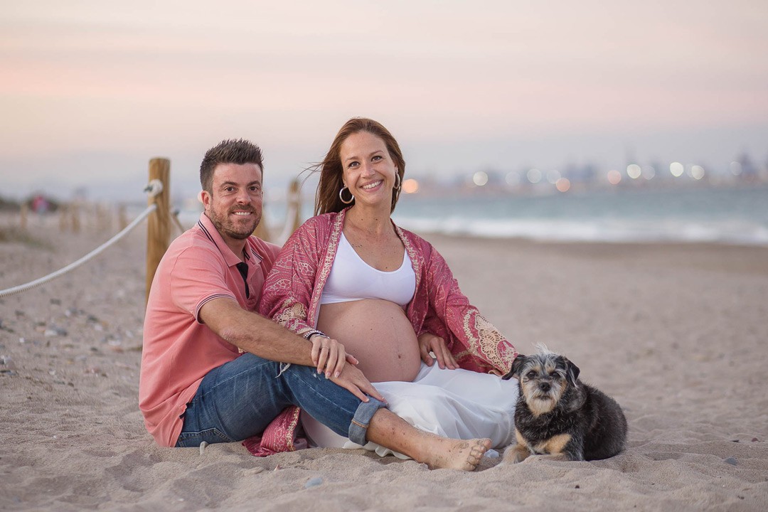 Chica embarazada junto a su pareja y su perro sentados en la playa