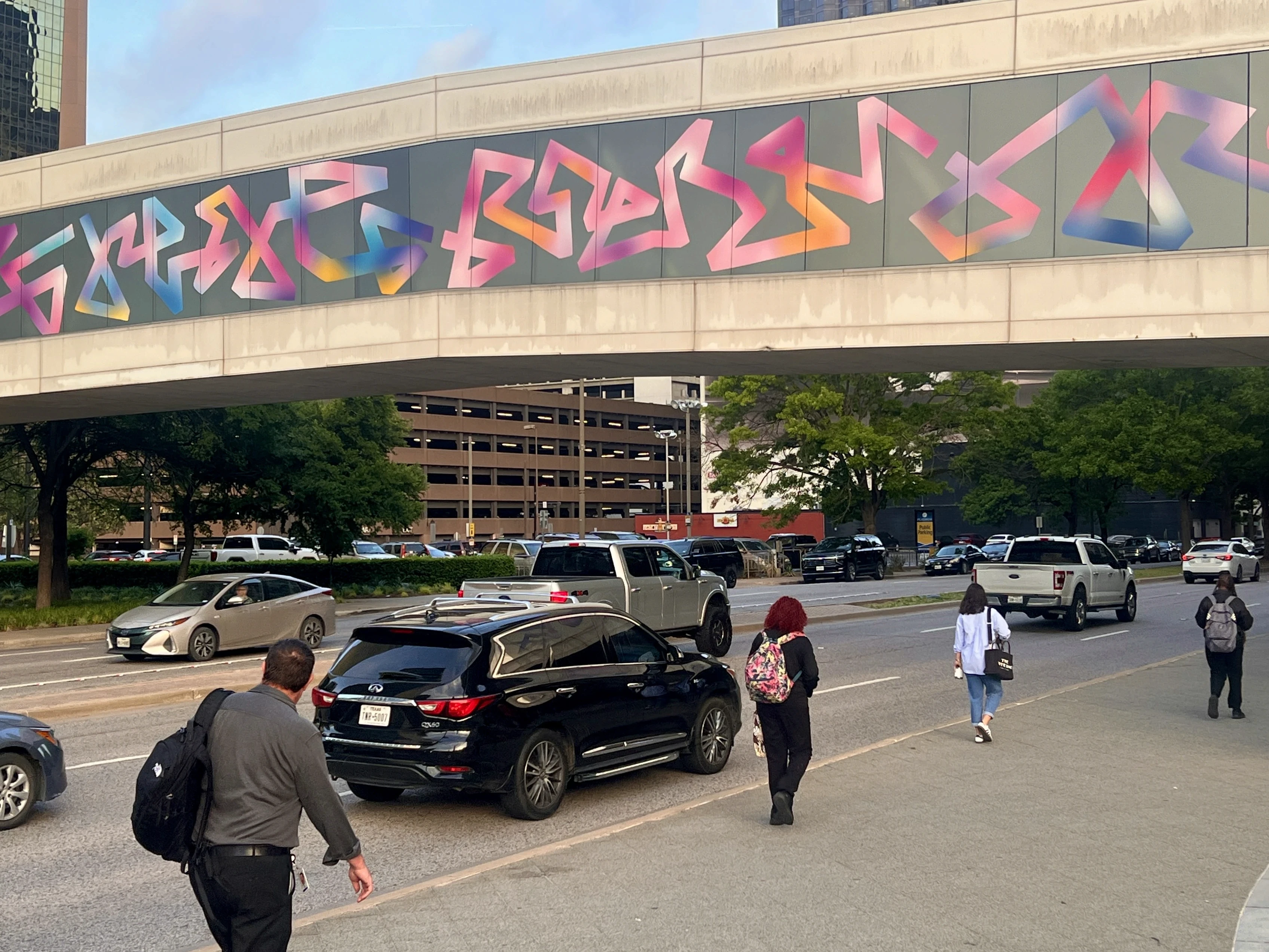 A busy city street with pedestrians and vehicles, featuring a colorful mural on a pedestrian bridge above.