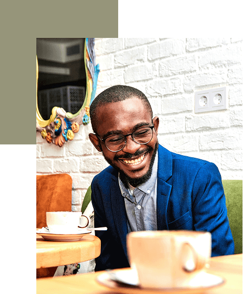 Man in a blue suit and glasses smiling while sitting at a café table with coffee cups, with a decorative mirror and white brick wall in the background.