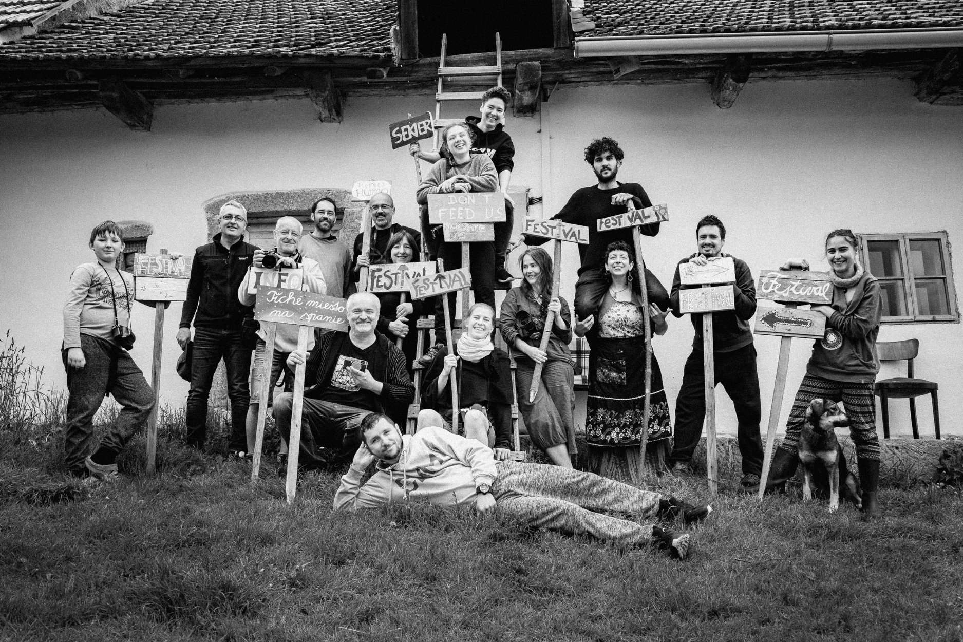 A young person with curly hair smiles at the camera while sitting in front of a laptop against a dark background.