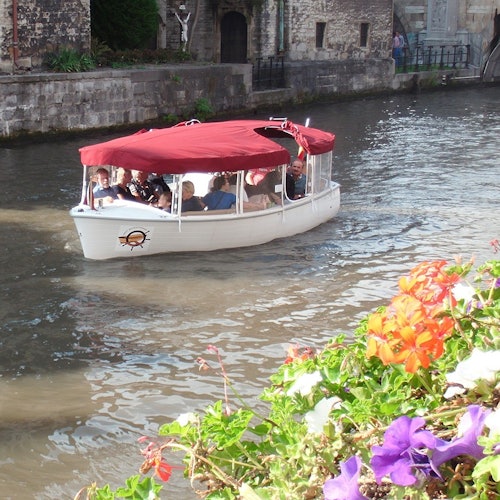 A boat with a red canopy carrying passengers moves along a canal lined with stone buildings and vibrant flowers in the foreground.
