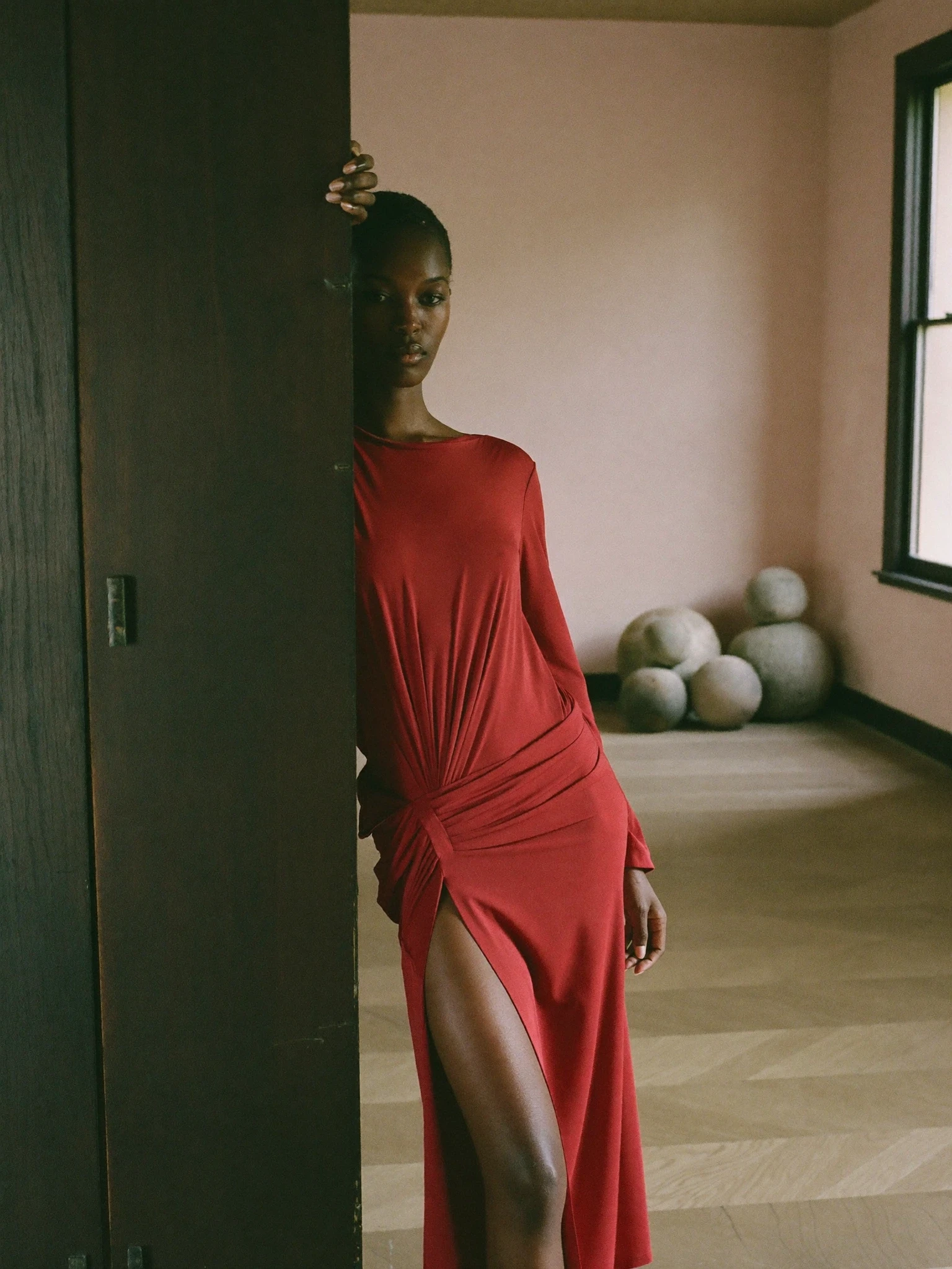 Model in a red draped dress with a high slit, leaning against a dark wood door frame