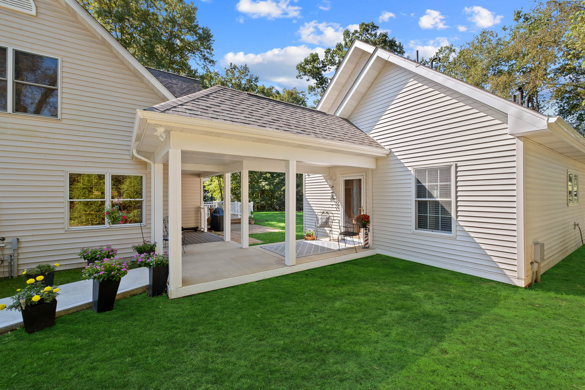 Rear exterior home addition with covered porch, white vinyl siding, gable roof, concrete patio, and landscaped backyard with green lawn and potted flowers.