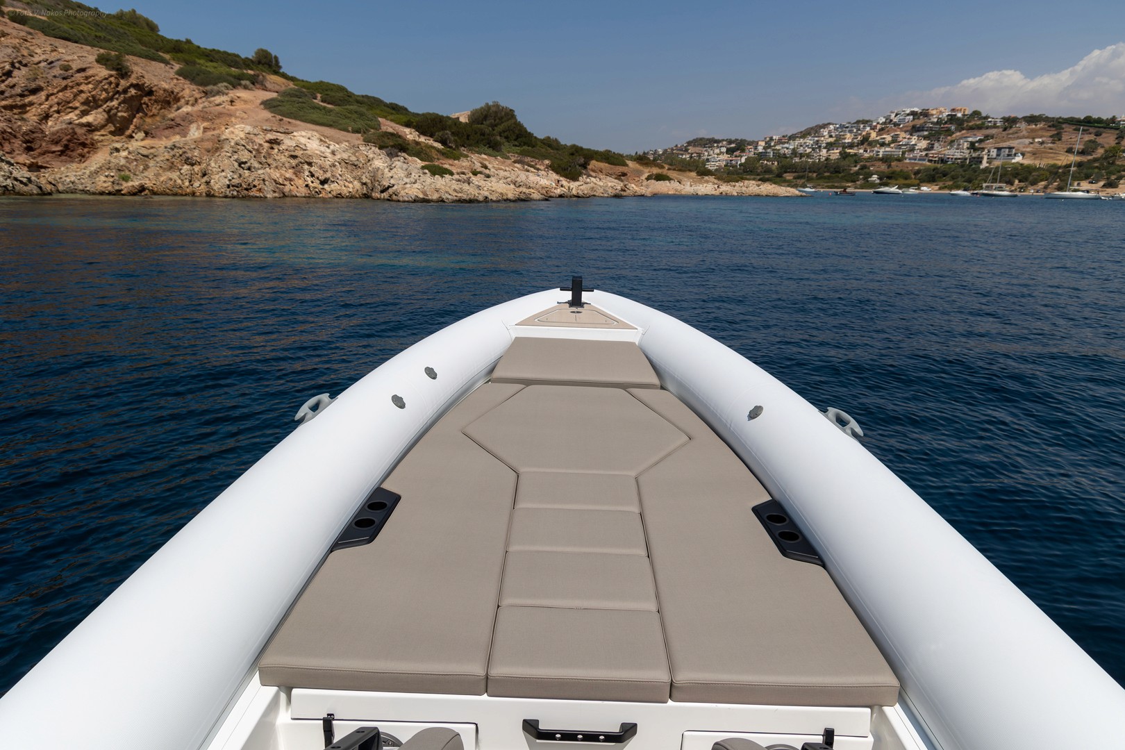 White and beige RIB boat bow with cushioned seating cruising through deep blue waters near rocky Greek coastline with hillside village.